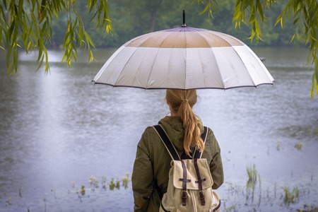 Young woman with umbrella looking at the rising river level during a flood. Rainy season. Female tourist with backpack in the rain.の写真素材