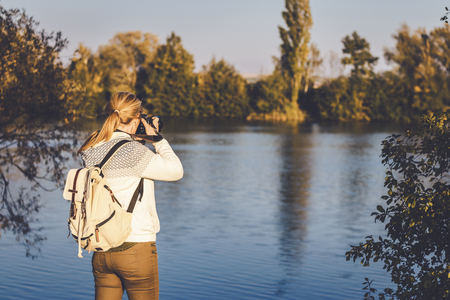 Woman with backpack is photographing a landscape near to lake, toned picture. Hiking woman in nature.の写真素材