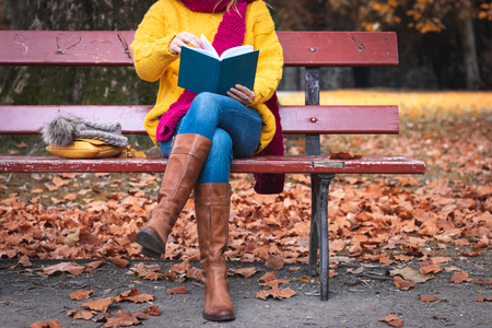 Woman sitting on bench and reading book. Autumn at public park. Woman wearing sweater, jeans and leather boots.の写真素材
