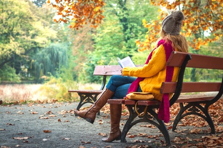 Woman sitting on bench and reading book. Autumn at public park. Woman wearing sweater, jeans and leather boots.の写真素材