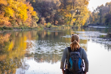 Woman is standing over a lake and looking to beautiful autumn landscape. Hiking in nature. Traveler with backpack.の写真素材