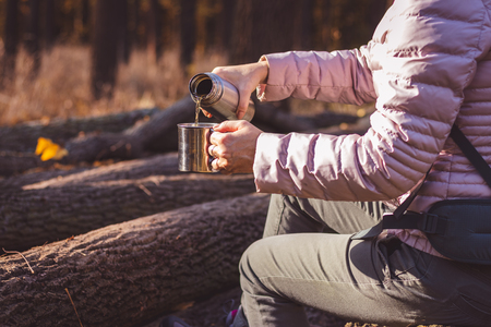 Woman pours hot tea out of thermos into metal cup in autumn forest. Refreshment during hiking in nature.の写真素材