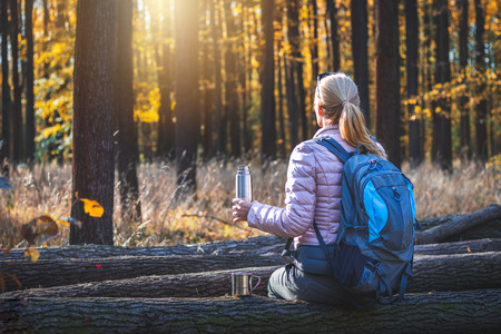 Relaxation in forest during autumn hiking. Female traveler with backpack holding a thermosの写真素材