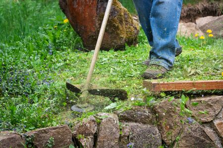 Gardener is mowing grass by brushcutter in garden near stone wall. Cutting grass at springtime.の写真素材