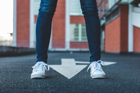 Woman wearing jeans and white sports shoe standing on asphalt road with directional sign. Slim female legs with sneakers in city streetの写真素材