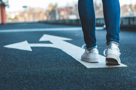 Make decision which way to go. Walking on directional sign on asphalt road. Female legs wearing jeans and white sneakers.