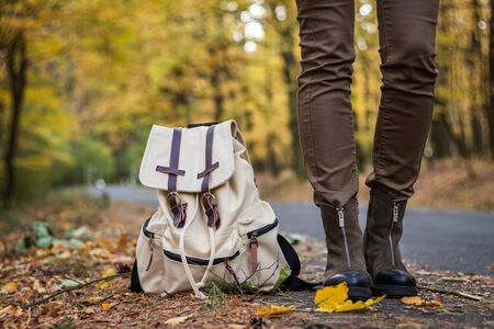 Woman with backpack wearing hiking boots is standing on road in autumn forest. Travel concept. Hitchhiker standing on roadsideの写真素材
