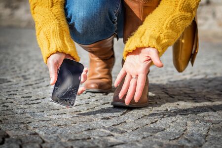 Woman feeling frustration from broken smartphone. Cracked touch screen on mobile phone. Woman picking up damaged smart phone on the streetの写真素材