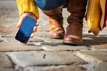 Woman picking up broken smartphone with cracked touch screen on the street. Damaged fragile device screen on mobile phone.の写真素材