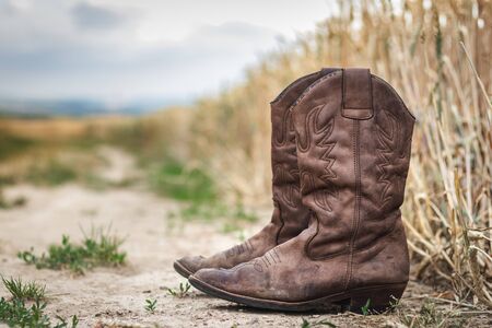 Cowboy boot on dirt road next to wheat field. Old worn out leather shoe outdoors. Retro style still life of footwear at rural sceneの写真素材