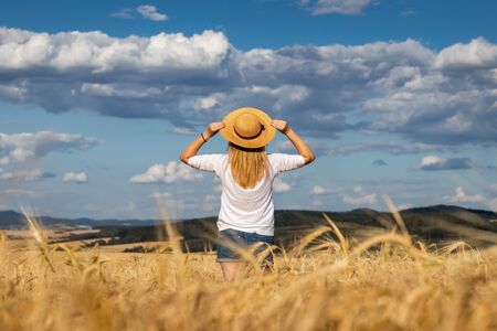 Happy woman with straw hat standing in wheat field and enjoying summer. Blond hair woman feeling freedom. Beautiful sky.の写真素材