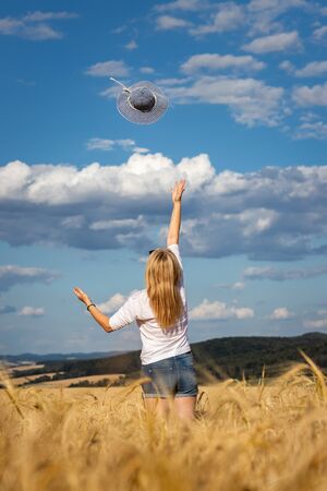 Happy blond hair woman throwing blue sun hat against beautiful sky. Enjoyment of summer. Carefree woman standing in wheat field.の写真素材