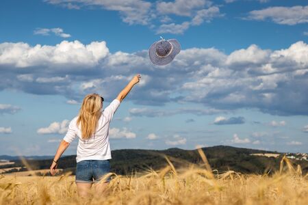 Happy blond hair woman throwing blue sun hat against beautiful sky. Enjoyment of summer. Carefree woman standing in wheat field.の写真素材