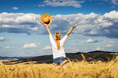 Happy woman holding straw hat with hand raised in wheat field. Enjoyment of summer. Carefree blond hair woman with arms outstretched against beautiful skyの写真素材