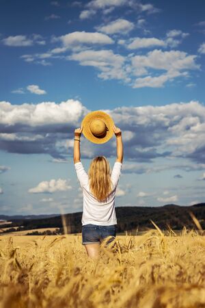 Happy woman holding straw hat is standing in wheat field. Enjoyment of summer. Carefree blond hair woman wearing casual clothing against beautiful skyの写真素材