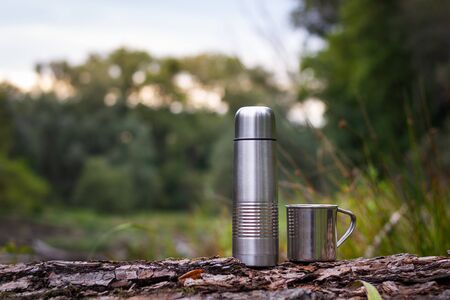 flask and stainless steel mug outdoors. Camping equipment on tree trunk in nature. Metal bottle and cup for hot drinkの写真素材