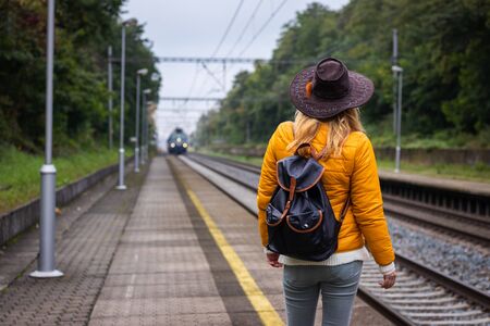 Woman with backpack and hat is looking at arriving train at railroad station platform. Traveler at beginnings of her journeyの写真素材