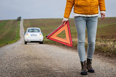Safety first. Broken car on the road. Driver holding warning triangle. Misfortune on travel. Woman need roadside assistance after car accidentの写真素材