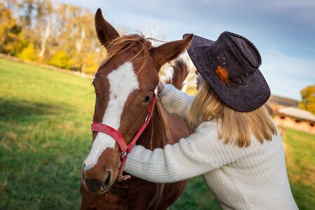 Woman with cowboy hat is hugging her young horse at pasture. Friendship between people and domestic animals. Cowgirl love her foalの写真素材