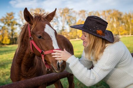 Woman playing with her young horse at pasture. Cowgirl love her foal. Friendship between people and domestic animals. Woman wearing cowboy hat and knitted sweater. の写真素材