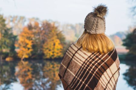 Woman with blanket and knit hat with pom-pom enjoying cold misty morning at lake. Enjoyment of autumn nature. Blond hair female wearing warm clothingの写真素材