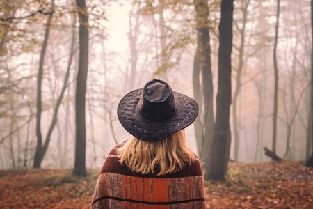Woman with hat wrapped in blanket is looking to fog. Moody atmosphere in misty forest at autumn. Enjoyment of nature in cold morningの写真素材