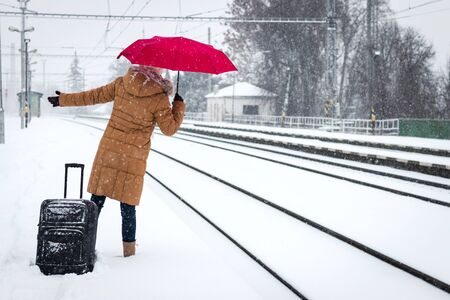 Where is the train? Delayed train during snow calamity. Woman with red umbrella and luggage waiting at railroad station in winter snowfallの写真素材