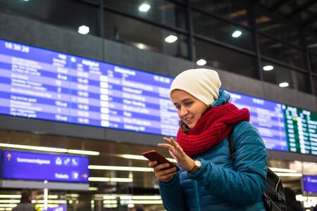 Young woman is booking railway ticket by using mobile app on her smartphone. Tourist is standing under arrival departure board or timetable at railroad stationの写真素材