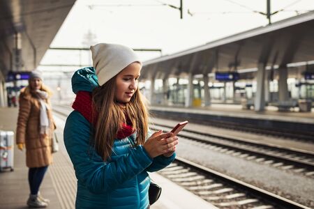 Woman is standing at railroad station and buying a railway ticket on her smart phone. People waiting for train on platform. Tourist using mobile app for booking ticket reservationの写真素材