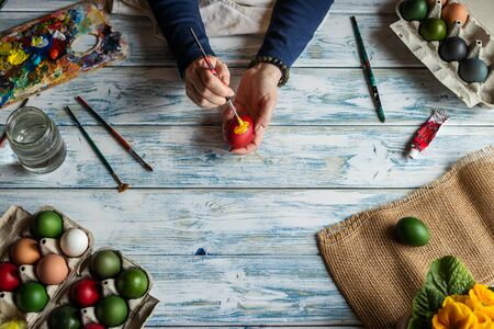Hands painting Easter eggs on white wooden table, top view. Woman coloring egg for traditional religious celebration. Artist using colors, paintbrush and palette for homemade decoration.の写真素材