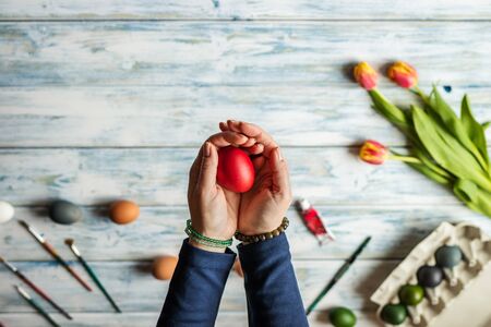 Woman holding coloring easter egg in her hands. White wooden table with tulip flower. Decorating for...