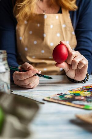 Woman wearing apron and coloring Easter eggs for traditional religious holiday. Red egg in female hand. Artist is ready for painting homemade decorationの写真素材