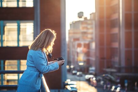 Woman photographing street at city. Blond hair woman standing at terrace and holding camera. Female tourist in European cityの写真素材