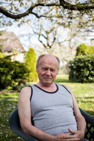 Portrait of happy senior man sitting in chair at his garden. Old man with smiling face relaxing in backyard at spring. Looking at cameraの写真素材