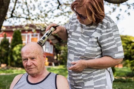 Woman is doing haircut with hair clipper to senior man outdoors at garden. Elderly people take care...