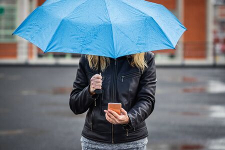 Woman with umbrella holding smartphone and standing in rain at street. Wireless internet connection on mobile phone in city の写真素材