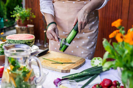 Woman peeling cucumber on table. Making fresh vegetable salad for garden party. Preparation of healthy vegetarian food outdoorsの写真素材
