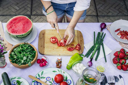 Preparing food on table outdoors. Woman cutting red tomato with kitchen knife on wooden board. Top viewの写真素材