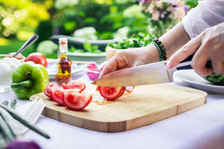 Woman cutting red tomato with kitchen knife on wooden board. Preparing food outdoors. Making fresh vegetable salad at gardenの写真素材