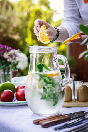 Sparkling water in carafe on table. Woman hand putting slice of orange into cold mineral water with mint and iceの写真素材