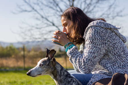 Woman drinking coffee with her whippet dog. Relaxation outdoors. Pet owner enjoying sunny morning with her cute dog at backyardの写真素材