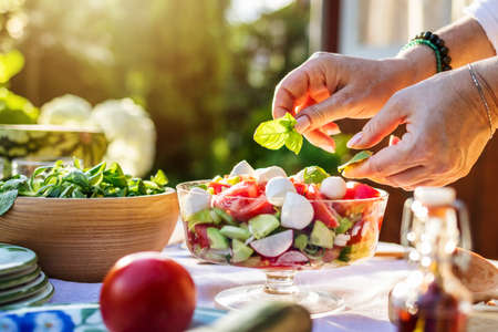 Female hand finishing fresh vegetable salad with herb basil leaves. Preparing healthy food in garden outdoorsの写真素材