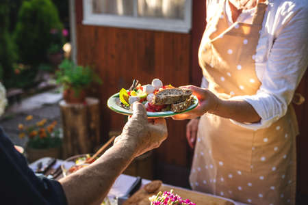 Woman serving vegetable salad with baguette on plate to another person at garden party. Catering service at celebration event outdoorsの写真素材