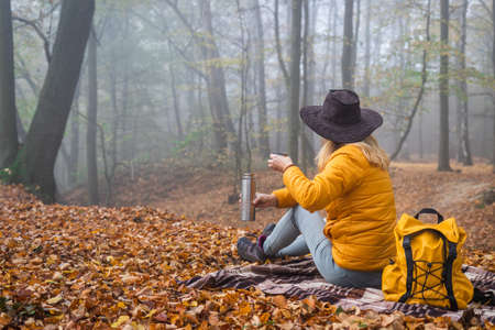 Hiker sitting on picnic blanket and drinking hot drink in autumn forest. Woman resting in nature during hikeの写真素材
