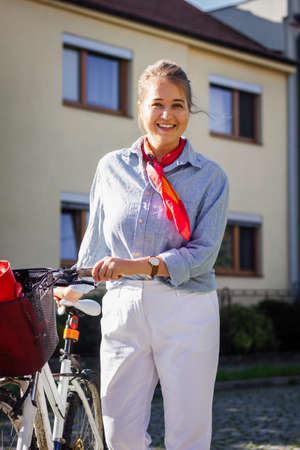 Smiling girl with bicycle wearing school uniform at city street. Beautiful young woman walking bike outdoorsの写真素材