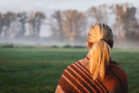 Woman with blanket standing in nature while camping outdoors. Cold autumn morning with fogの写真素材