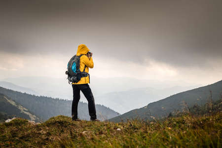 Adventure in nature. Hiker photographing mountain range in bad weather. Woman with backpack hiking outdoors. Tourist taking photograph during hikeの写真素材