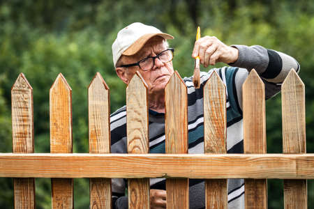 Senior man painting wooden fence in garden. Renovation of old wood picket fence. Domestic life and work in retirementの写真素材