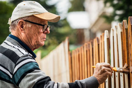 Senior man painting wooden fence at backyard. Old man paint picket fence by wood stainの写真素材