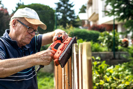 Sander in hands. Senior man sanding wooden fence in garden. Old carpenter using electric work toolの写真素材